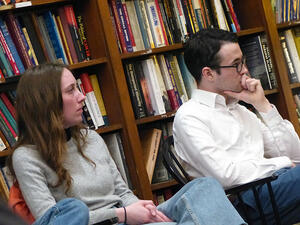 Two students sit in chairs in a library and listen to a speaker