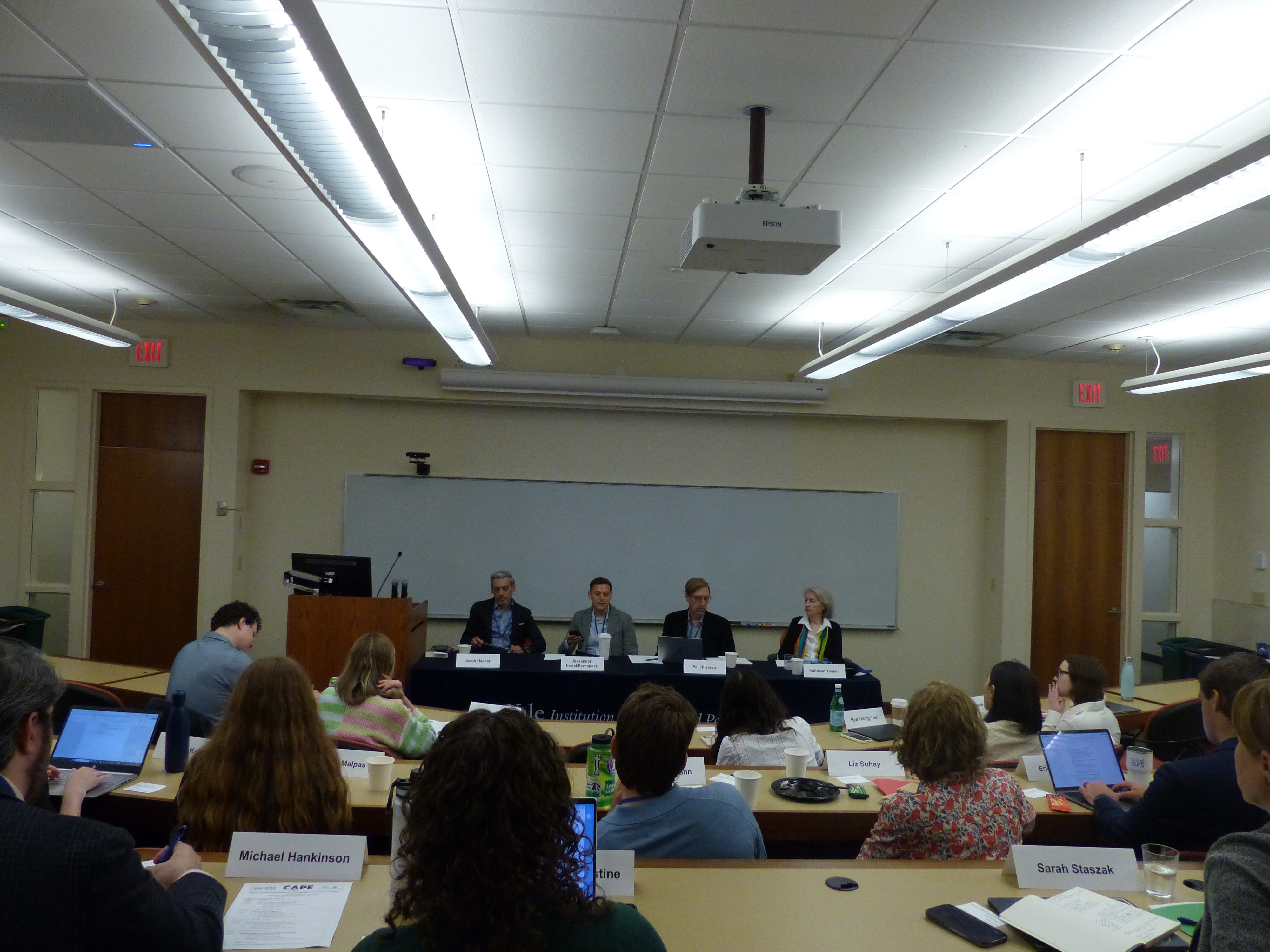 Panelists sit at a table at the front of a classroom, as seen from a seat in back