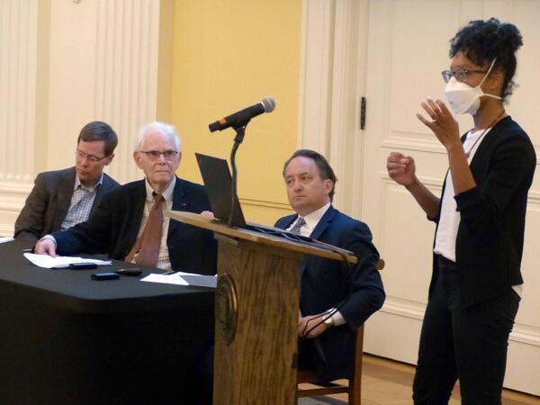Allison Harris speaks at a lectern as Greg Huber, David Mayhew, and Pericles Lewis listen at a table