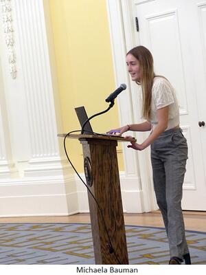 Michaela Bauman speaks in a round yellow room from behind a lectern