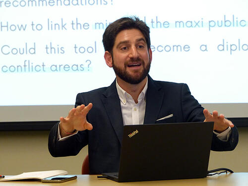 Jonathan Moskovic speaks at a table in a classroom with a projector screen behind him