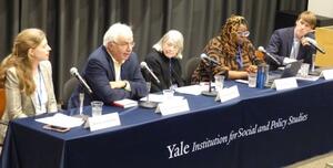 Five panelists sit behind a blue table in an auditorium, speaking into microphones