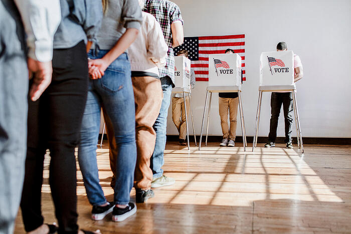 Voters wait in line in front of three voting booths, with an American flag on the wall