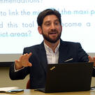Jonathan Moskovic speaks at a table in a classroom with a projection screen behind him