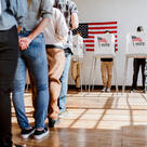 Voters wait in line in front of three voting booths, with an American flag on the wall