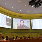 Attendees of a session in a circular classroom listen to Kevin DeLuca speak from one of several large screens high on the walls.